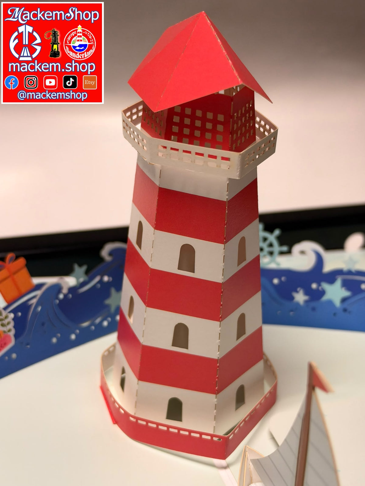 A red and white striped paper pop-up lighthouse model displayed on a white surface with a decorative blue and white nautical background. A red and white logo overlay for 'Mackemshop' is visible in the top-left corner.
