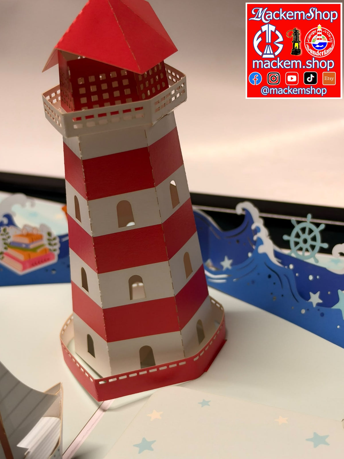 A 3D papercraft pop-up lighthouse with red and white horizontal stripes, displayed on a decorated pop-up book page with blue waves and a ship wheel.