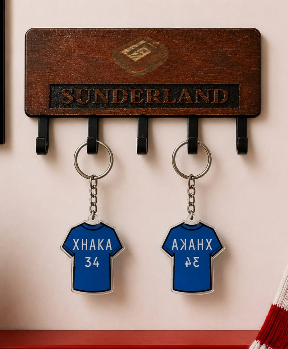 A wooden Sunderland football club key holder mounted on a wall, featuring two blue jersey-shaped keychains with player names and numbers. A Sunderland scarf, framed poster, and potted plant are visible in the styled setting.