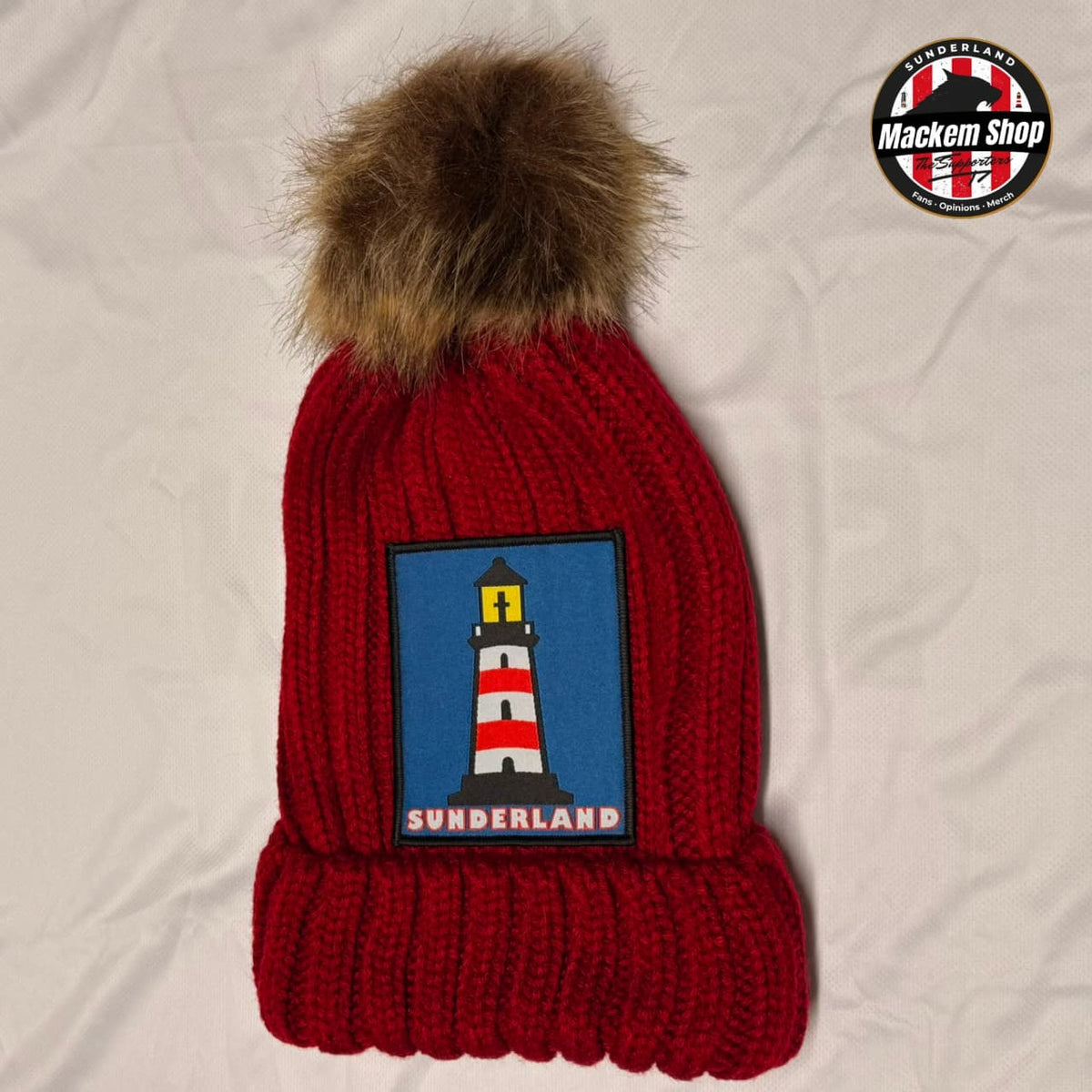 A red knit beanie with a brown faux fur pom-pom and a blue patch featuring a lighthouse and the word 'SUNDERLAND'. A circular 'Mackem Shop' logo is visible in the top right corner.