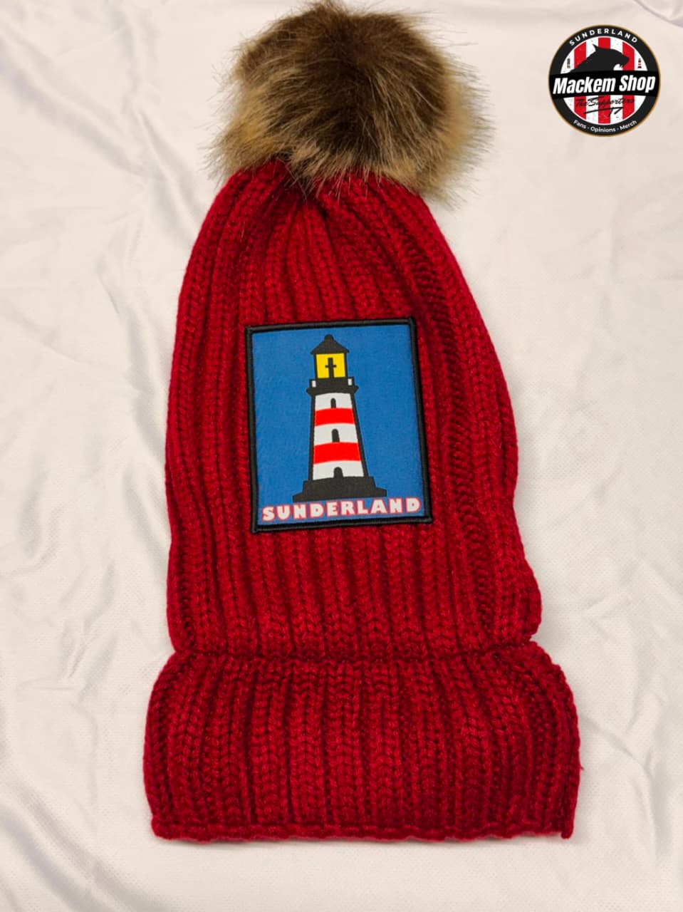 A red ribbed knit beanie with a brown faux-fur pom pom, featuring a rectangular patch with a lighthouse and the word 'SUNDERLAND' on a blue background. A circular 'Mackem Shop' logo is visible in the top right corner.