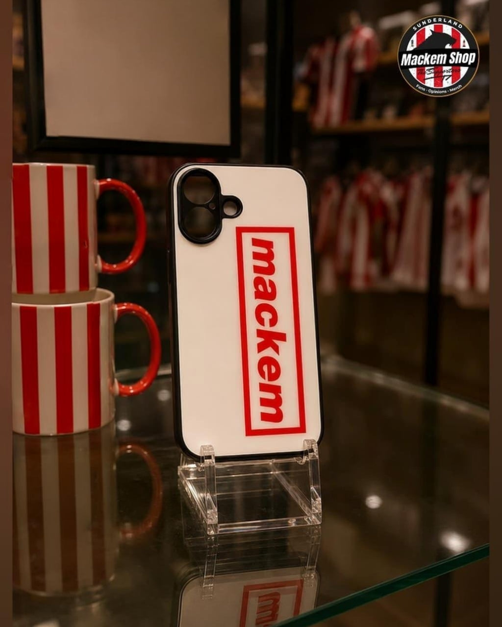 A white phone case with a red 'mackem' logo displayed on a clear stand on a glass shelf, with red and white striped mugs in the background.
