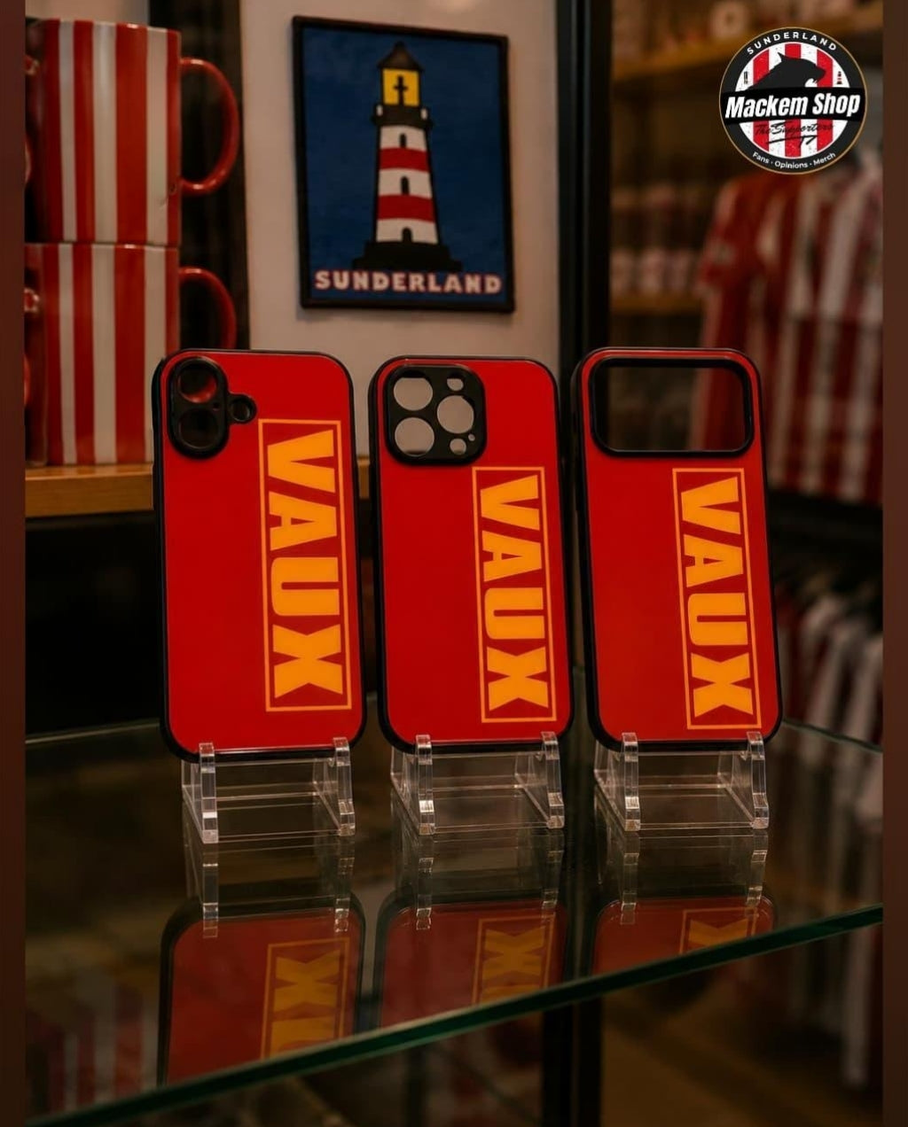 Three red phone cases with bold yellow 'VAUX' lettering displayed on clear stands on a glass shelf, with a Mackem Shop logo in the top right corner.
