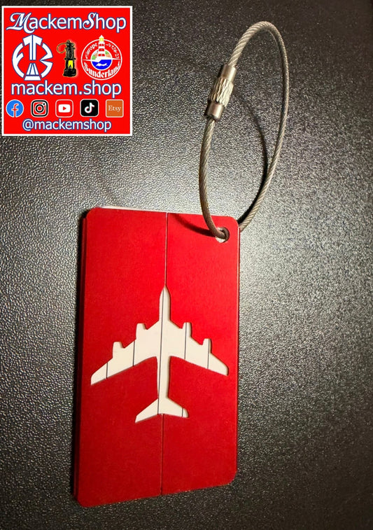 A red luggage tag with a white airplane silhouette, attached to a gray metal cable, shown on a dark textured background. A 'Mackemshop' logo overlay is in the top-left corner.