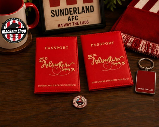 Two red passport-style clutch bags with gold 'Adventure' lettering and Sunderland AFC text, displayed on a wooden table with a Sunderland AFC sign, a red mug, a scarf, a round pin, and a keychain.