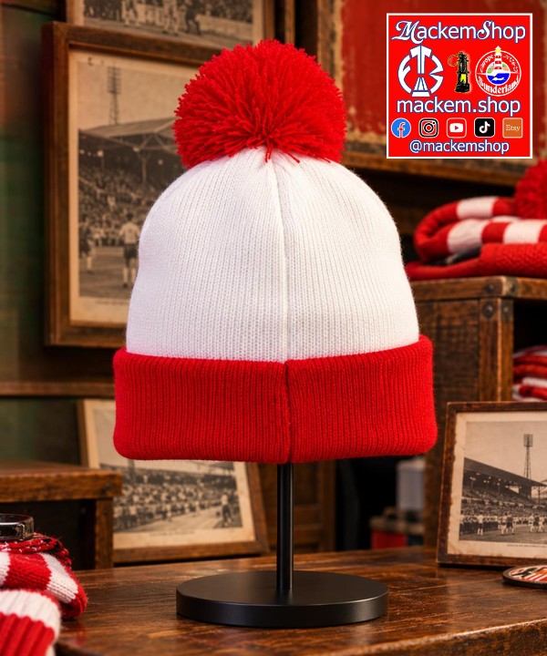 A red and white knit beanie with a large red pom-pom, displayed on a black stand in a rustic shop setting with framed football stadium photos and a red-and-white striped scarf nearby. A 'MackemShop' logo overlay is in the top right corner.
