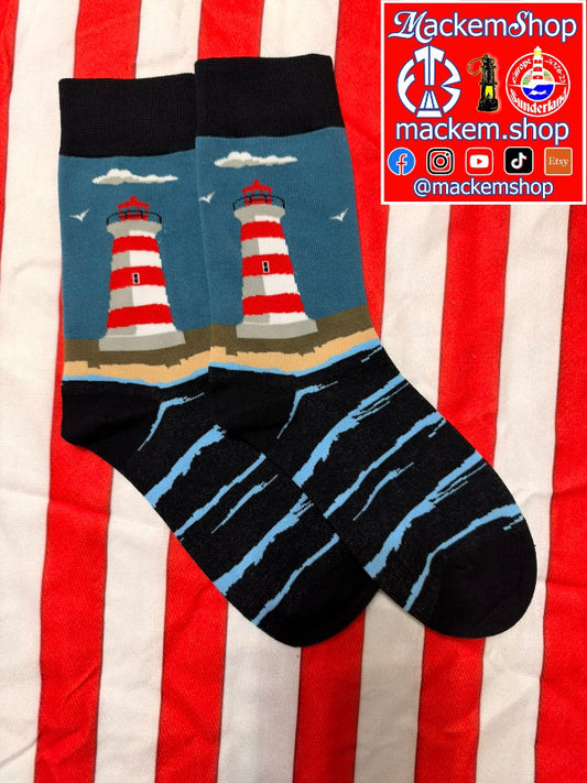 A pair of crew socks laid flat on a red and white striped fabric background. The socks have a dark blue upper section with two red-and-white striped lighthouses, white clouds, and a tan shoreline, while the lower portion is black with light blue abstract stripes, and a MackemShop logo on the top right corner.