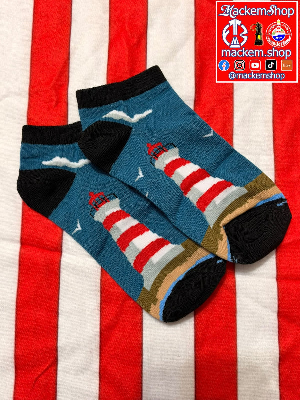 A pair of ankle socks featuring a lighthouse and seagulls design on a blue background, styled on a red and white striped fabric backdrop. A brand logo for 'MackemsSop' is visible in the top right corner.