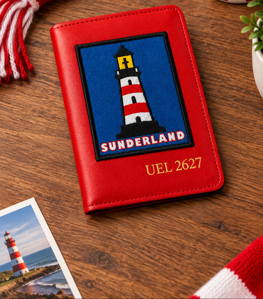 A red faux-leather passport cover featuring a Sunderland football club crest patch with a lighthouse on a blue background, and the text 'SUNDERLAND' and 'UEL 2627' in gold. The cover is displayed on a wooden surface with a blurred Sunderland scarf, a red Sunderland jersey, and a passport in the background.