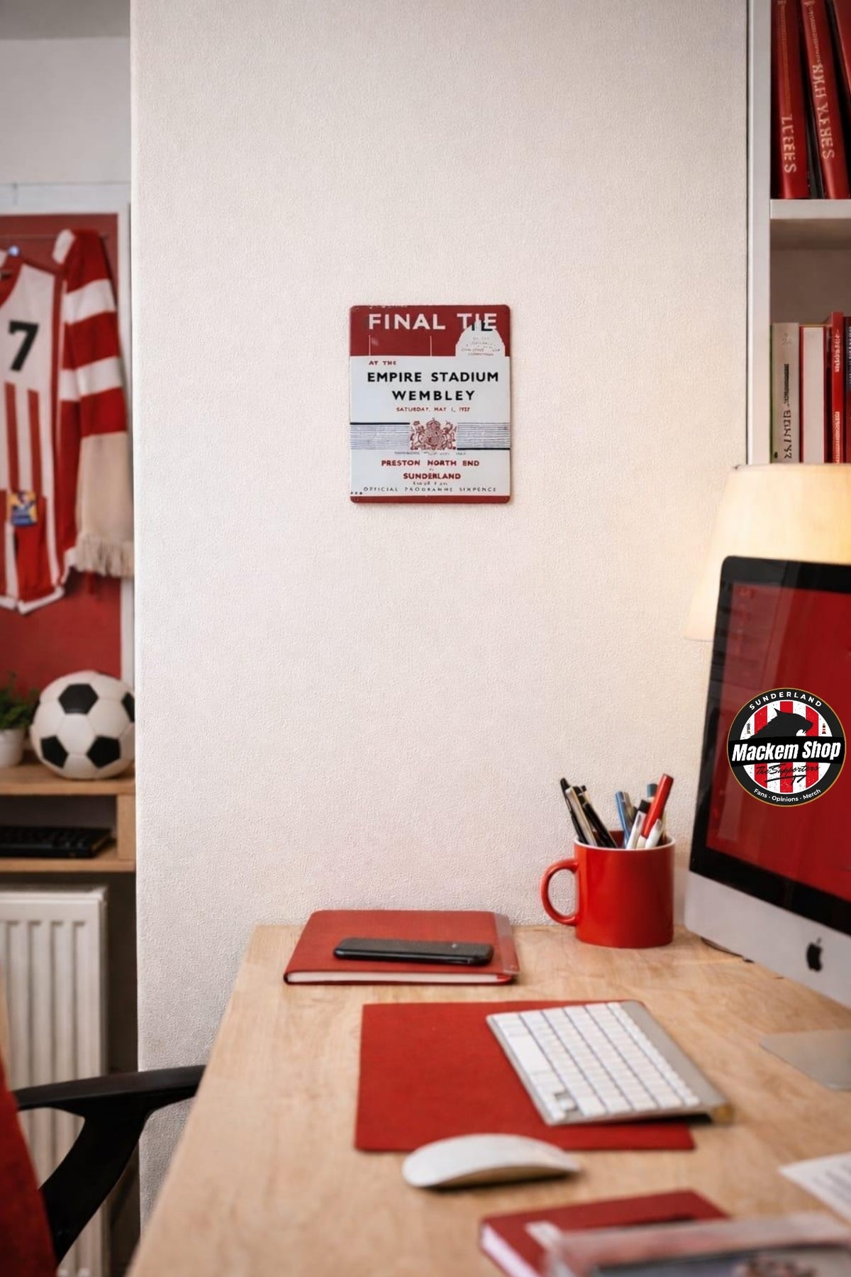 A red and white vintage-style Stadium Wembley sign mounted on a white wall in a home office setting. The desk below features a red notebook, a white keyboard, a red mug, and an iMac displaying MackemShop logo.