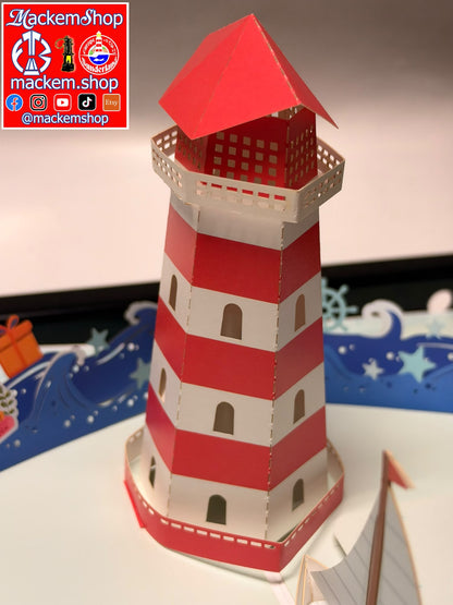 A red and white striped paper pop-up lighthouse model displayed on a white surface with a decorative blue and white nautical background. A red and white logo overlay for 'Mackemshop' is visible in the top-left corner.