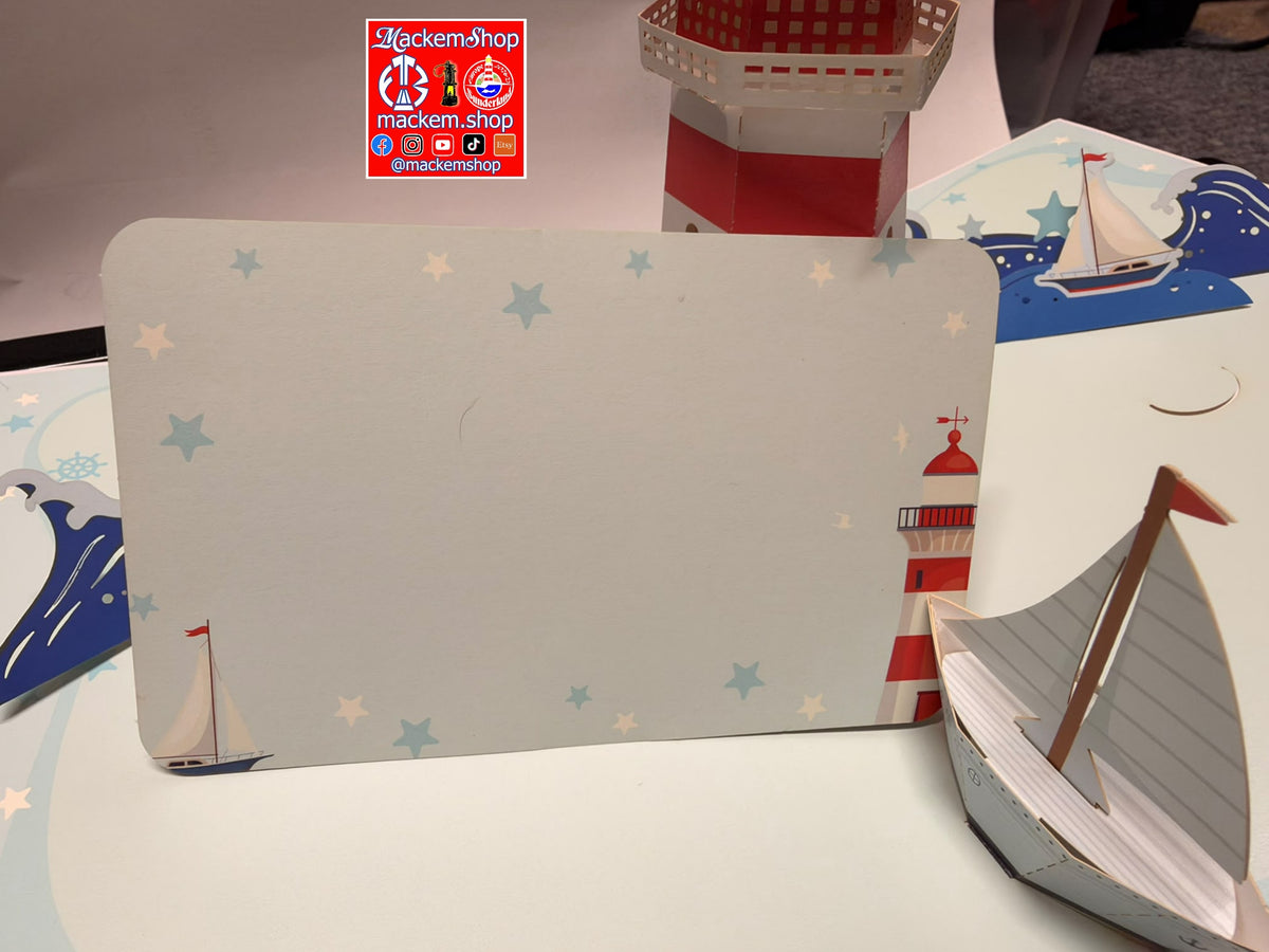 A handmade nautical-themed paper craft display featuring a large rectangular panel with a lighthouse and stars, a small paper boat in the foreground, and a red-and-white striped cup in the background. A 'Mackemshop' logo is visible on the table.