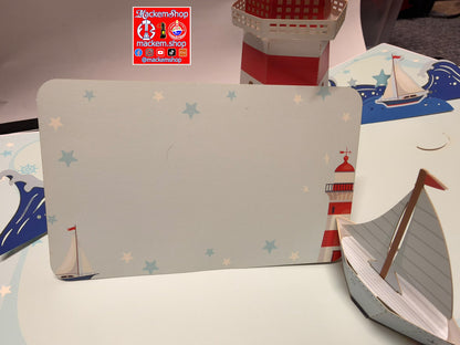 A handmade nautical-themed paper craft display featuring a large rectangular panel with a lighthouse and stars, a small paper boat in the foreground, and a red-and-white striped cup in the background. A 'Mackemshop' logo is visible on the table.