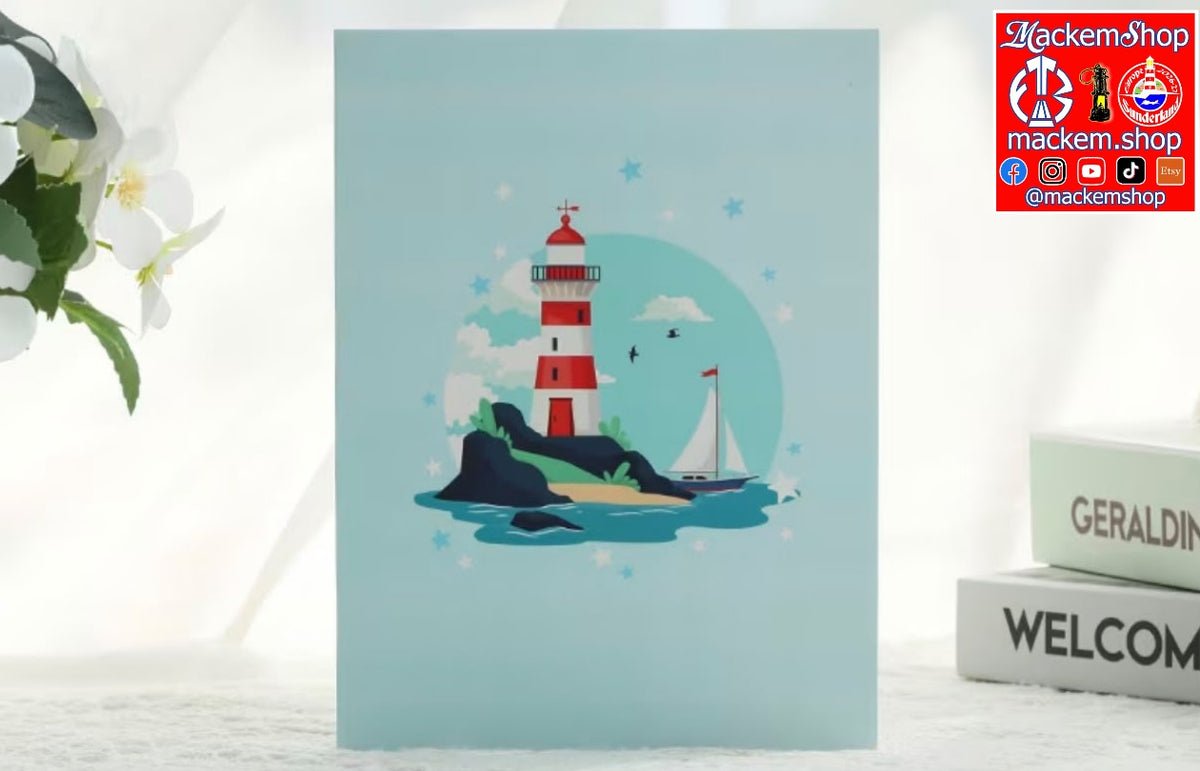 A light blue greeting card featuring a red and white striped lighthouse, a small sailboat, and a circular moon design, displayed on a white surface next to decorative books and flowers.