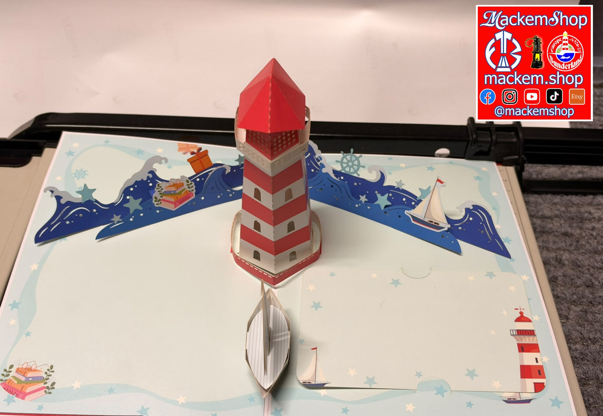 A 3D pop-up cardboard nautical scene featuring a red-and-white striped lighthouse, blue waves, sailboats, and a small boat in the foreground, displayed on a tabletop with a visible MackemShop logo overlay in the top right.