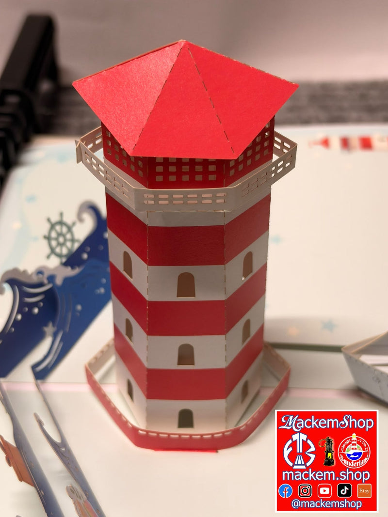 A red and white striped paper lighthouse model with a red pointed roof, displayed on a white surface with a blue ship-themed card in the background. A 'MackemShop' logo is visible in the bottom right corner.