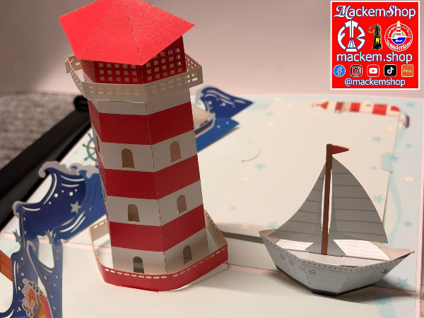 A 3D pop-up paper model set featuring a red and white striped lighthouse and a small paper sailboat, displayed on a decorated pop-up book page. A watermark for 'MackemShop' is visible in the top right corner.