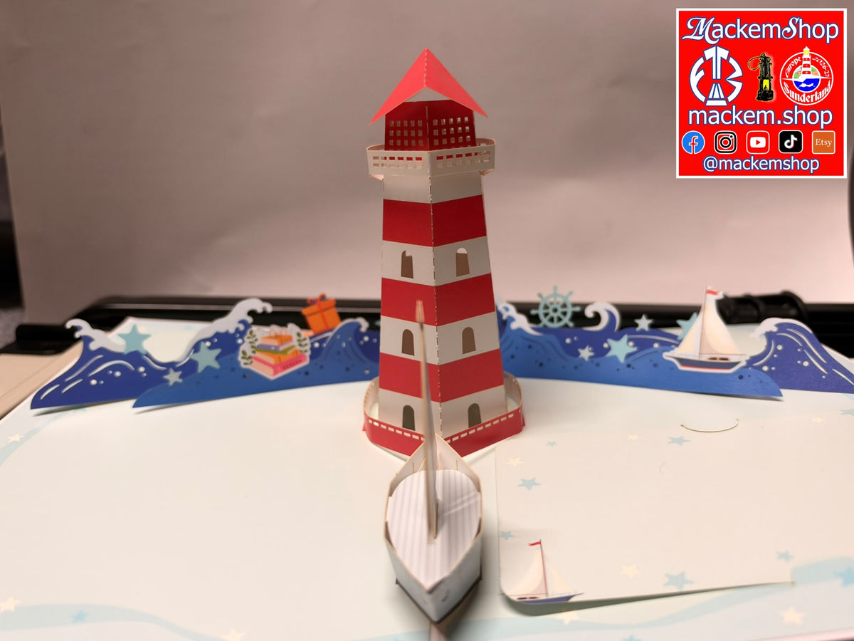A 3D papercraft pop-up model of a red and white striped lighthouse with small paper boats and blue ocean waves, displayed on a light gray surface. A Mackemshop logo sticker is visible in the top right corner.