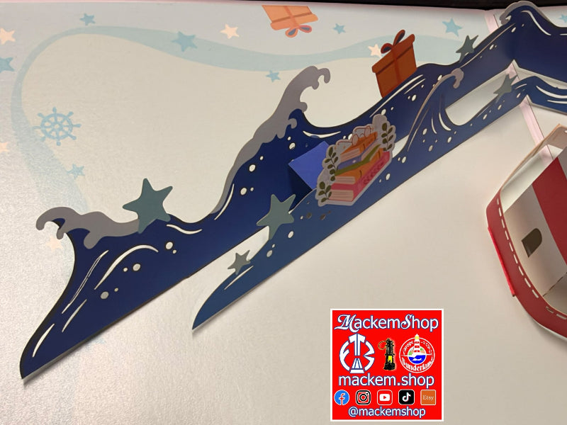 A close-up of a decorative paper craft or card featuring layered blue waves, gray stars, and a small gift box, photographed on a white surface with a visible MackemShop logo .