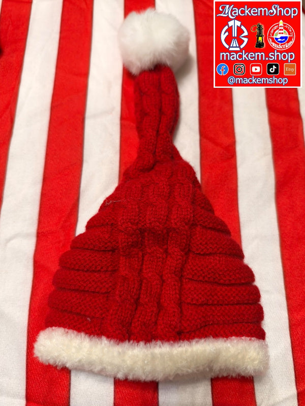 A red knitted Santa hat with a white faux-fur trim and pom-pom, laid flat on a red and white striped fabric background. A 'MackemShop' logo overlay is visible in the top right corner.