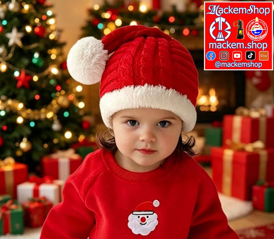 A toddler wearing a red knit Santa hat with a white pom-pom and a red Santa sweatshirt with a white Santa face graphic, posed in front of a decorated Christmas tree with wrapped gifts. A 'MackemShop' logo overlay is visible in the top right corner.
