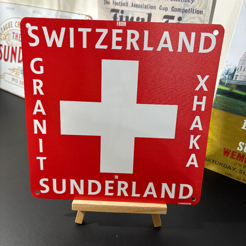 Red sign with white cross and Granit Xhaka text on a stand, with a blurred background of books and papers.