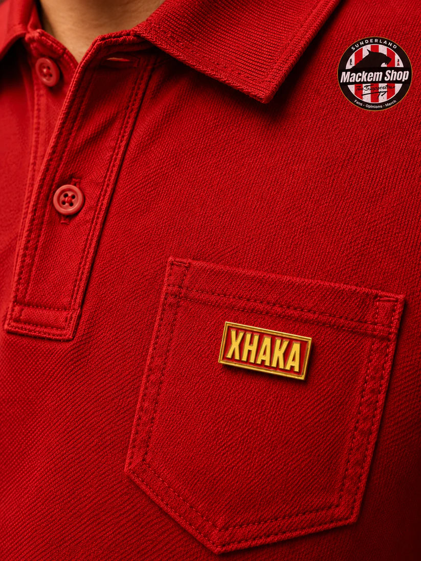 A close-up of a red polo shirt featuring a custom rectangular metal name patch with 'XHAKA' in gold lettering on the chest pocket. A circular 'Mackem Shop' brand logo is visible on the upper right chest.