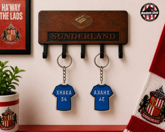 A wooden Sunderland football club key holder mounted on a wall, featuring two blue jersey-shaped keychains with player names and numbers. A Sunderland scarf, framed poster, and potted plant are visible in the styled setting.