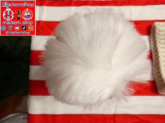 A fluffy white faux fur pom-pom hat or accessory shown from above on a red and white striped fabric background, with a knit cream item partially visible at the right edge with a MackemShop logo in the left corner.