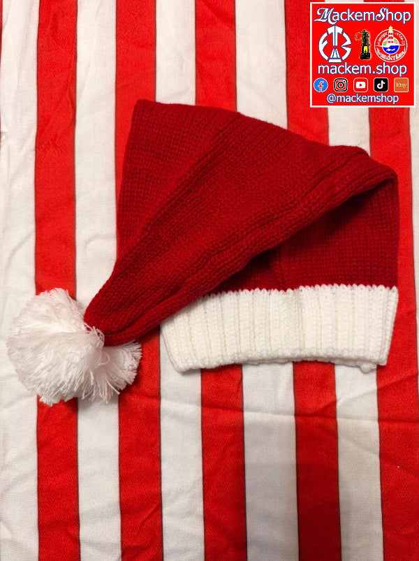 A red and white knitted Santa hat with a fluffy white pom-pom, laid flat on a red and white vertically striped fabric background. A MackemShop logo watermark is visible in the top right corner.