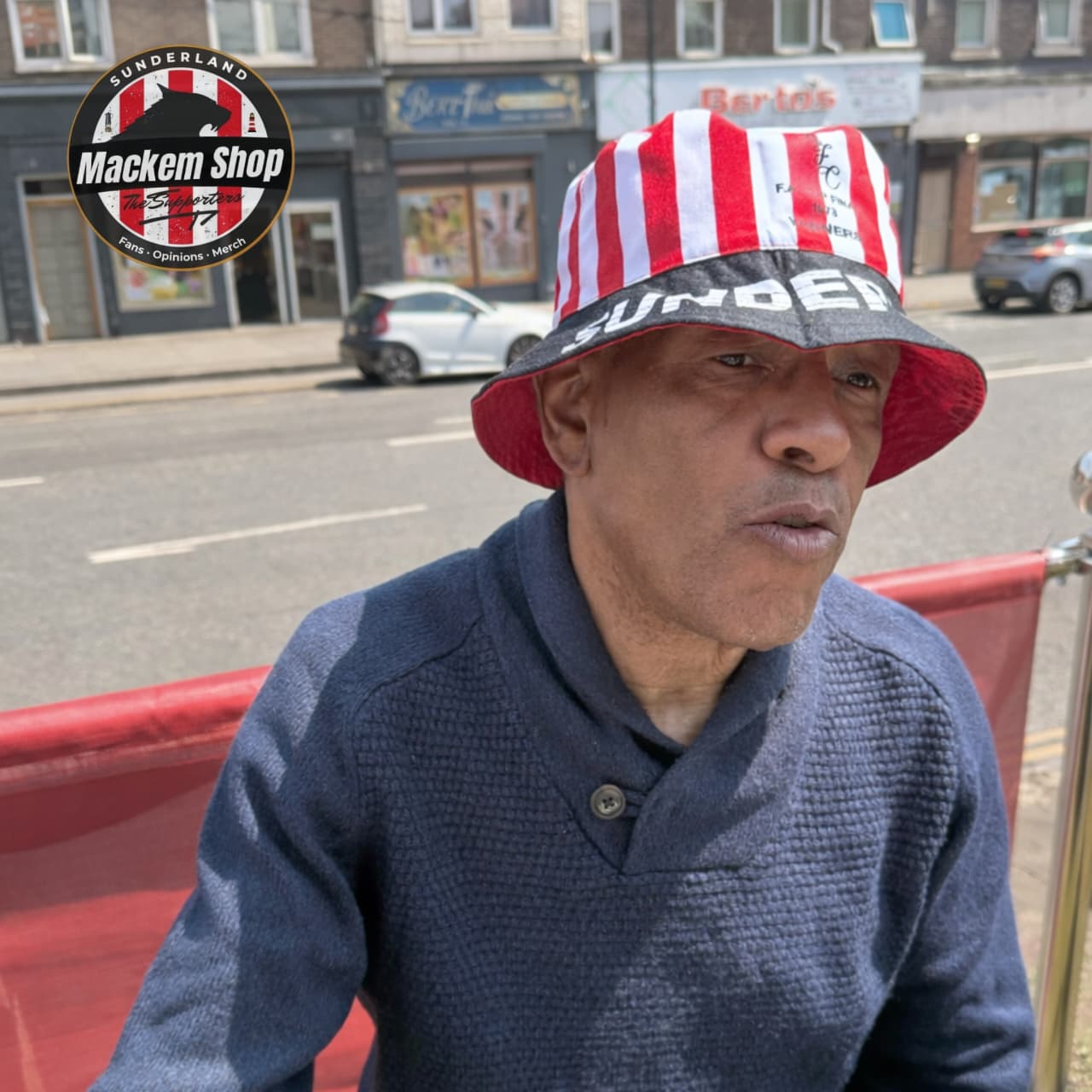 Man wearing a red and white striped bucket hat with 'Under' branding, sitting on a bench in an urban setting.