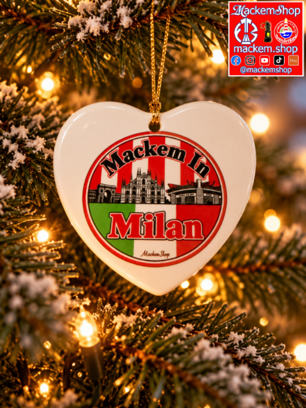 A heart-shaped white ceramic Christmas ornament hanging on a snowy Christmas tree with warm string lights. The ornament features a circular red, white, and green design with the text 'Mackem In Milan' and a small illustration of a building.
