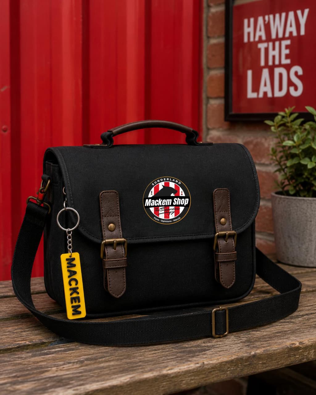 Black satchel bag with 'Mackem Shop' logo and keychain on a wooden surface with red wall and plant in the background.