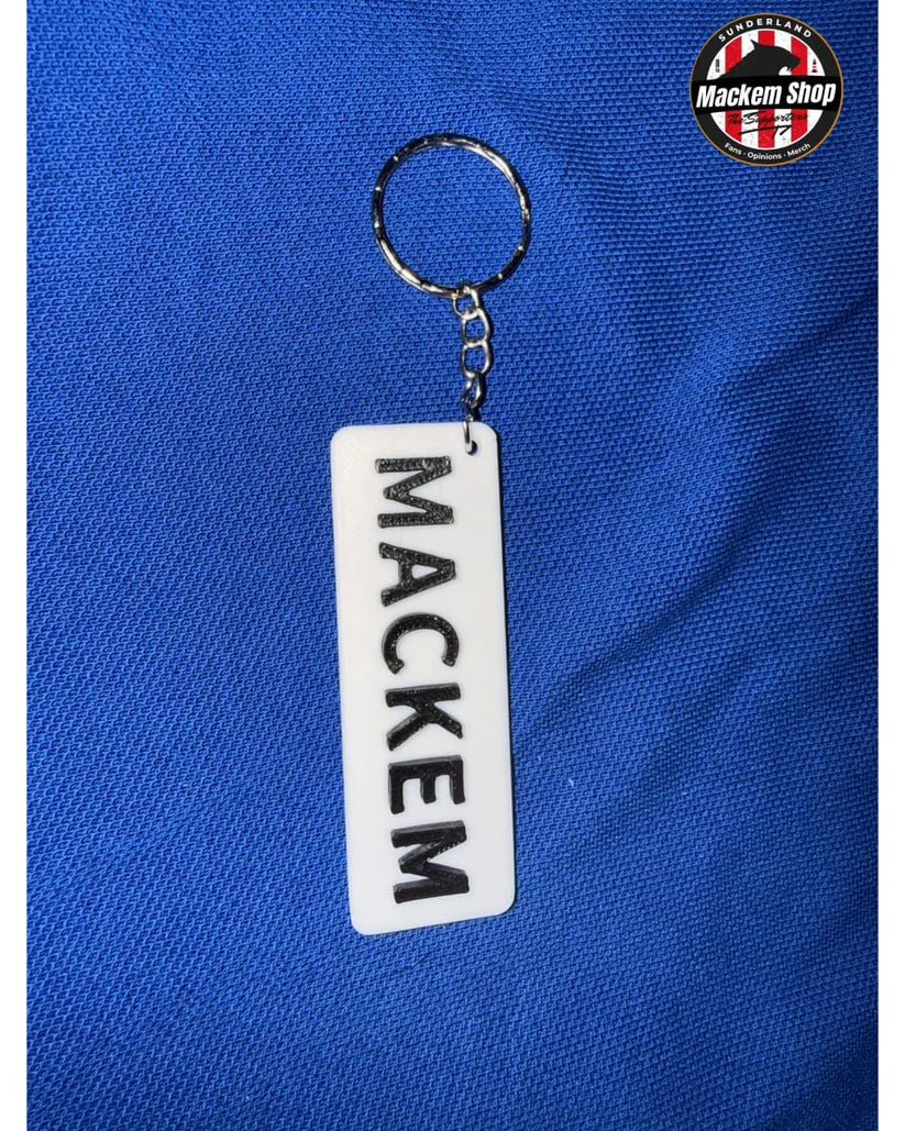 A white rectangular acrylic keychain with the name 'MACKEM' printed vertically in black, attached to a silver metal key ring, displayed on a textured blue fabric background. A circular 'Mackem Shop' logo is visible in the top right corner.