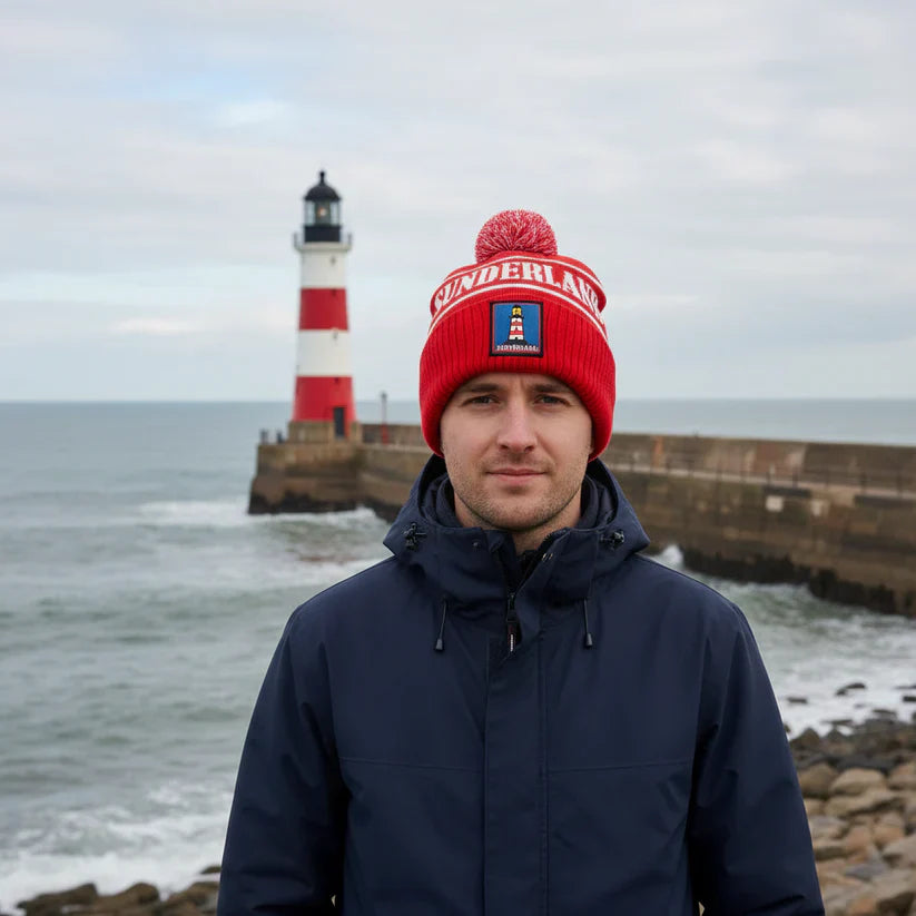Man wearing a red beanie and dark jacket standing in front of a lighthouse on a rocky shore.