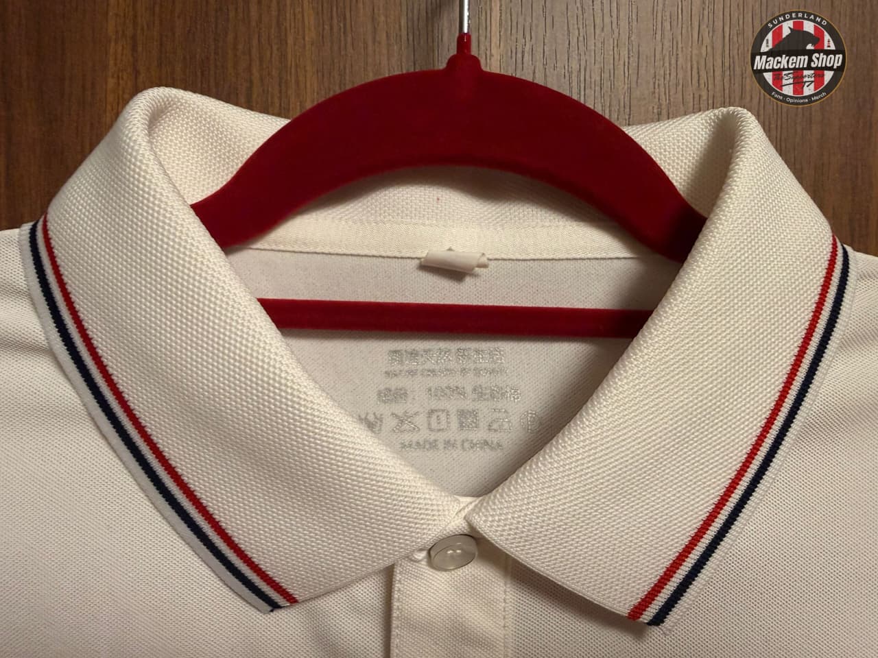 White polo shirt with red, blue, and black striped collar on a hanger against a wooden background.