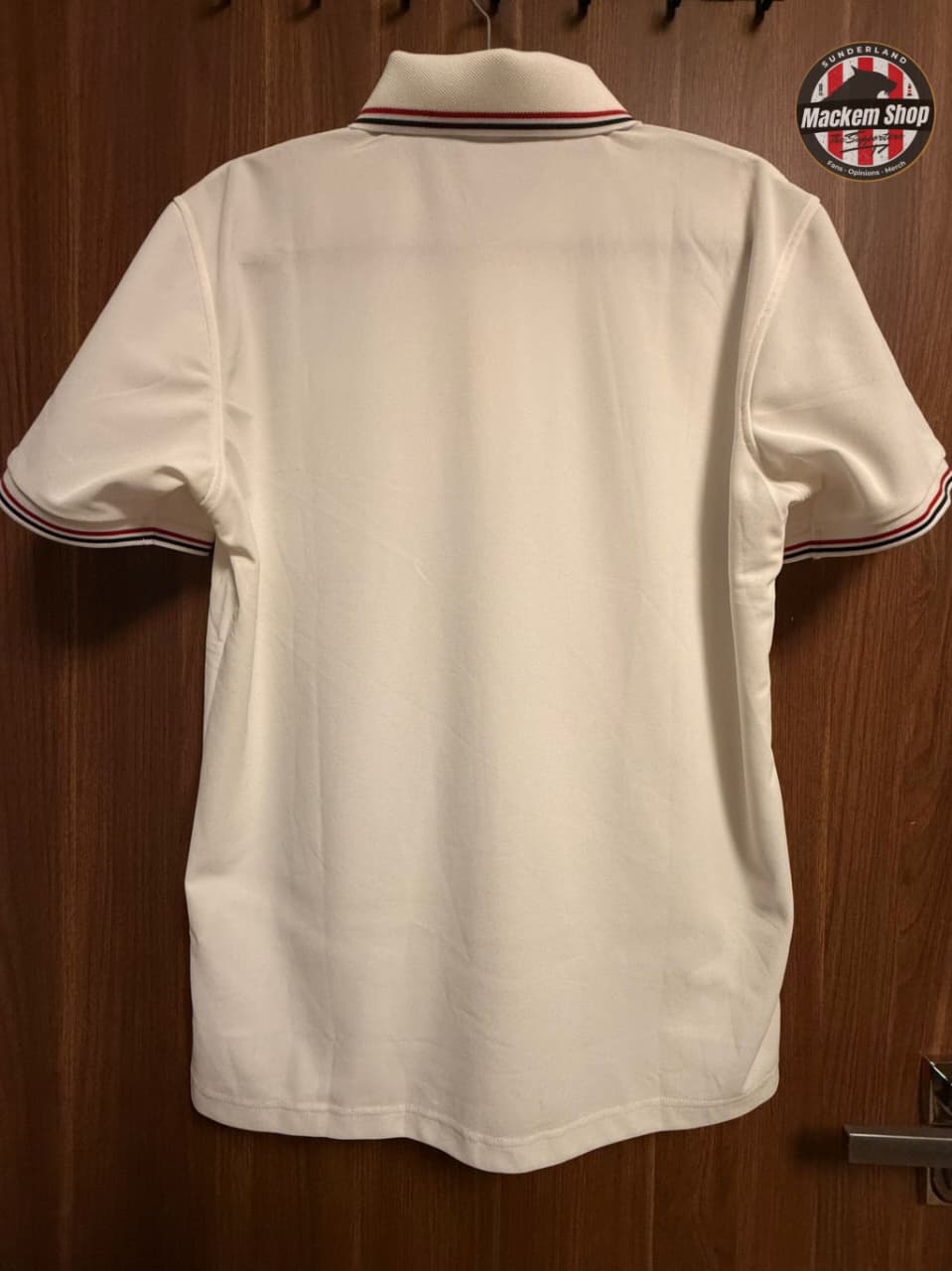 White polo shirt with red and blue trim on a wooden surface, branded 'Mackem Shop'.