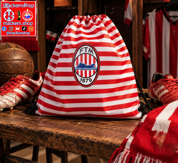 A red and white striped drawstring bag with a central FTM 1879 crest, displayed on a wooden bench in a sports-themed room with soccer shoes and a scarf.