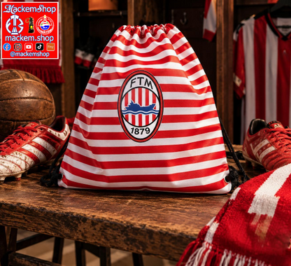A red and white striped drawstring bag with a central FTM 1879 crest, displayed on a wooden bench in a sports-themed room with soccer shoes and a scarf.