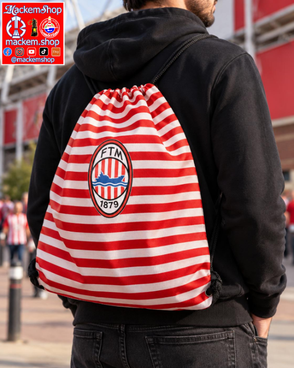 A man viewed from behind wearing a red and white striped drawstring backpack with the FTM 1879 crest on the back. The man is wearing a black hoodie and dark jeans, standing outdoors in front of a stadium.