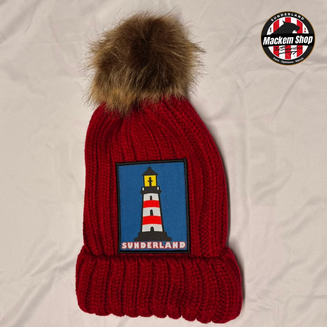 A red knit beanie with a brown faux fur pom-pom and a blue patch featuring a lighthouse and the word 'SUNDERLAND'. A circular 'Mackem Shop' logo is visible in the top right corner.