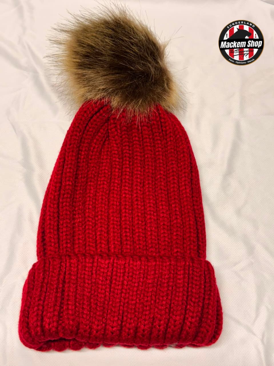 A bright red ribbed knit beanie with a large brown faux fur pom-pom, laid flat on a white fabric background. A circular 'Mackem Shop' logo is visible in the top right corner.
