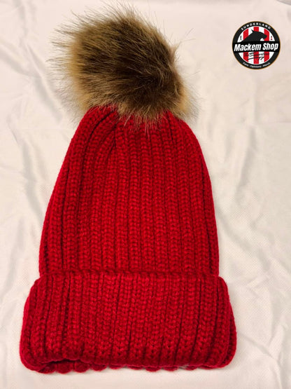A bright red ribbed knit beanie with a large brown faux fur pom-pom, laid flat on a white fabric background. A circular 'Mackem Shop' logo is visible in the top right corner.