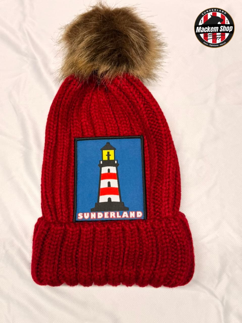 A red ribbed knit beanie with a brown faux-fur pom-pom, featuring a square patch of the Sunderland cityscape with a lighthouse on a blue background. A circular 'Mackem Shop' logo is visible in the top right corner.