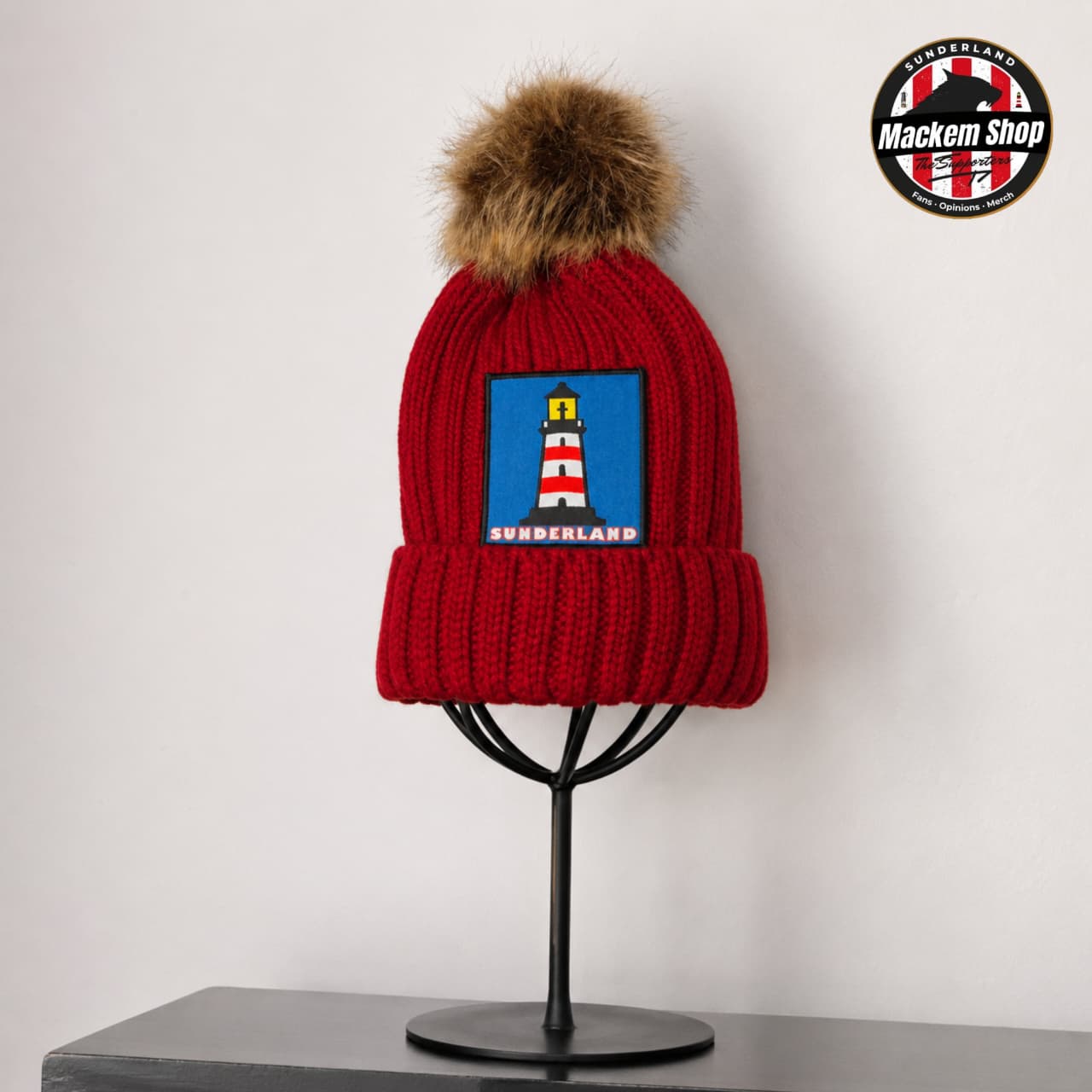 A red ribbed knit beanie with a brown faux-fur pom-pom and a blue patch featuring a lighthouse and the text 'SURRENLAND', displayed on a black stand against a light gray wall. A circular 'Mackem Shop' logo is visible in the top right corner.