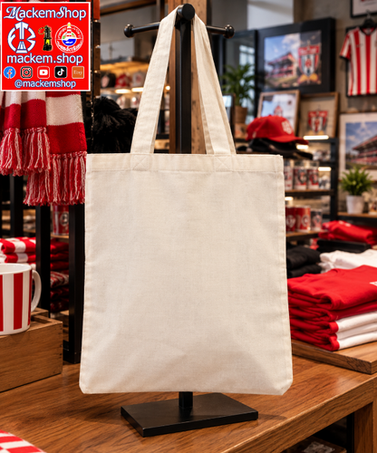An off-white canvas tote bag with two shoulder straps, displayed on a black stand inside a retail store. The background shows sports merchandise, including a red and white striped scarf, folded red and white shirts, and framed pictures.