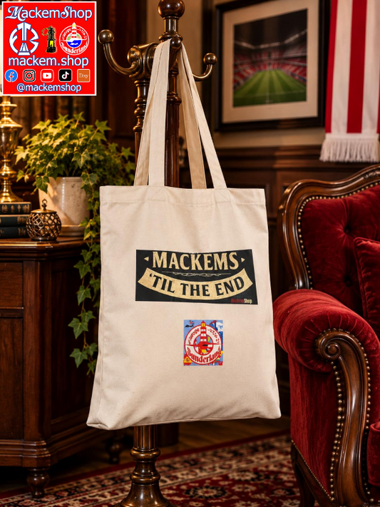 A cream canvas tote bag with 'MACKEMS TIL THE END' and a small circular logo printed on the front, hanging from a wooden coat rack in a vintage-style room with a red velvet armchair and framed wall art, with MackemShop Logo visible on left corner.