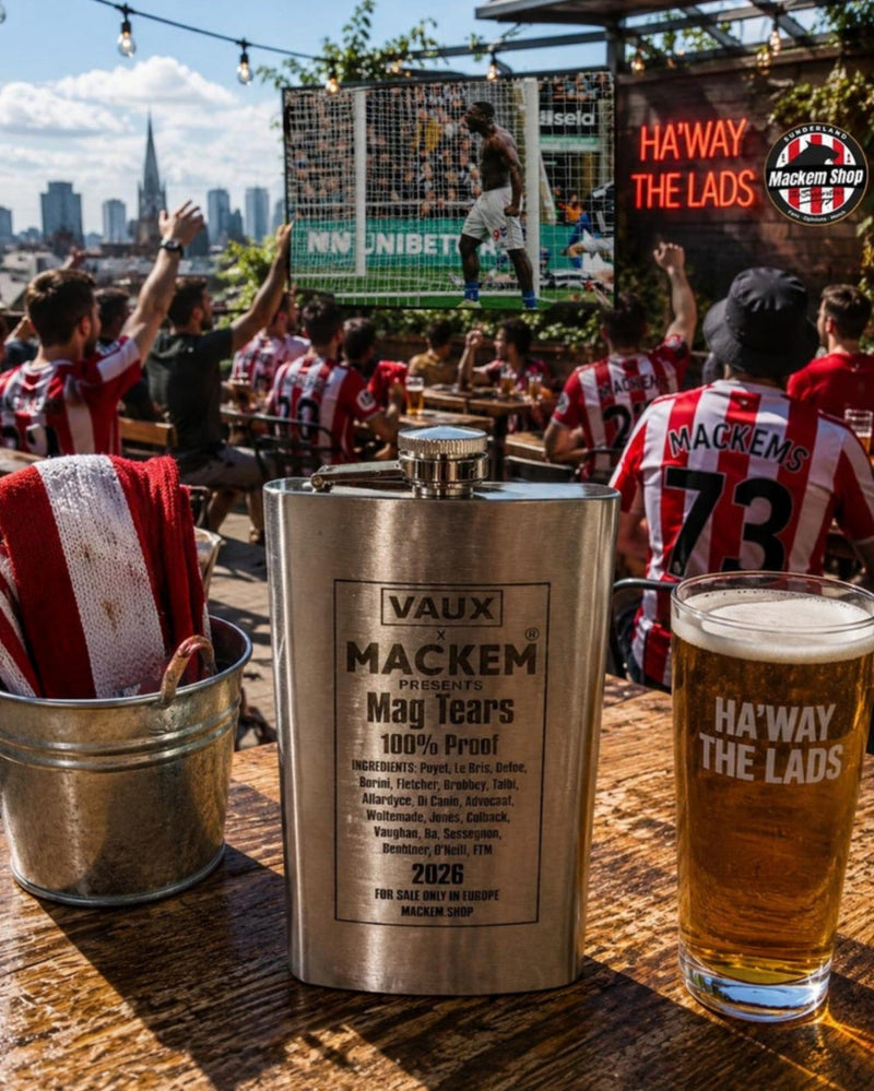 A stainless steel Vaux Mackem mag tear flask sits on a wooden bar table next to a glass of beer and a bucket of red and white striped napkins, with a lively sports crowd in the background.