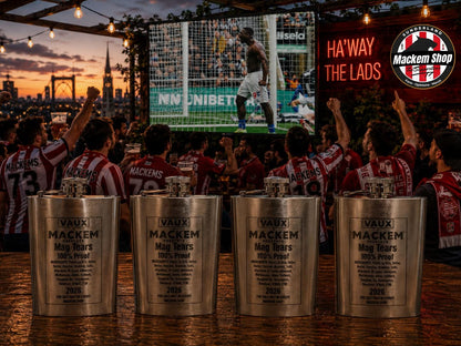 Four engraved silver Vaux Mackem mag tears Flask displayed on a bar counter in a crowded outdoor bar scene at sunset, with soccer fans in red and white striped jerseys celebrating in the background.