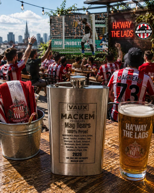 A silver Vaux Mackem Mag Tears flask sits on a wooden table at an outdoor bar, surrounded by people wearing red and white striped jerseys, a pint of beer, and a bucket of red towels. A large screen behind shows a soccer game.